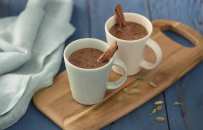 Fotografia em tons de azul em uma bancada de madeira azul, um pano azul, uma tábua de madeira com duas xícaras brancas com o chocolate quente com canela dentro delas.